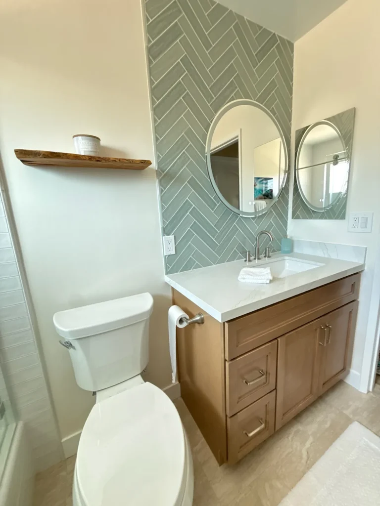 Wood vanity with round mirrors and a floating shelf against green herringbone tile